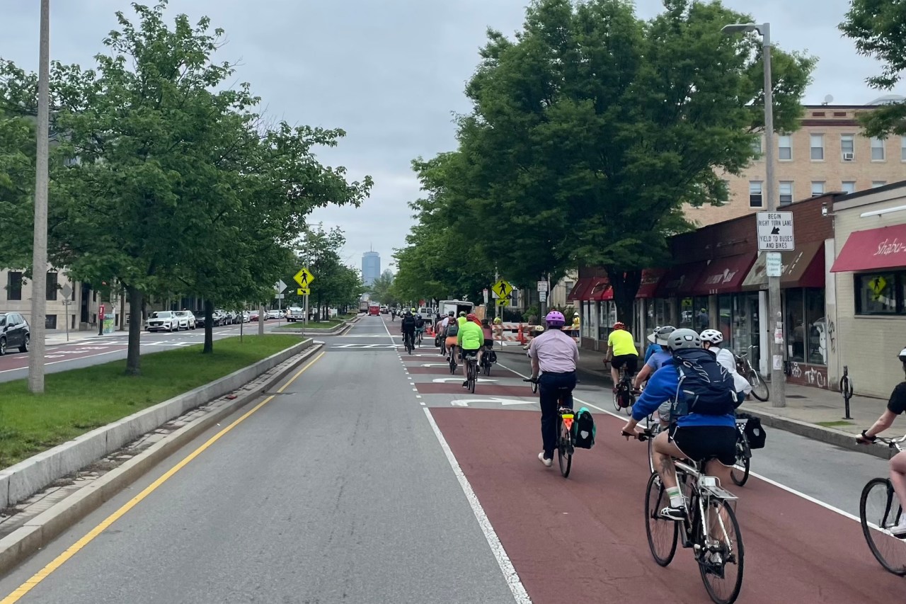 A crowd of people on bikes ride one- and two-abreast along a red-painted bus and bike lane along a multi-lane city street lined with trees. In the distance, a single skyscaper, the Prudential Center, is visible on the horizon.
