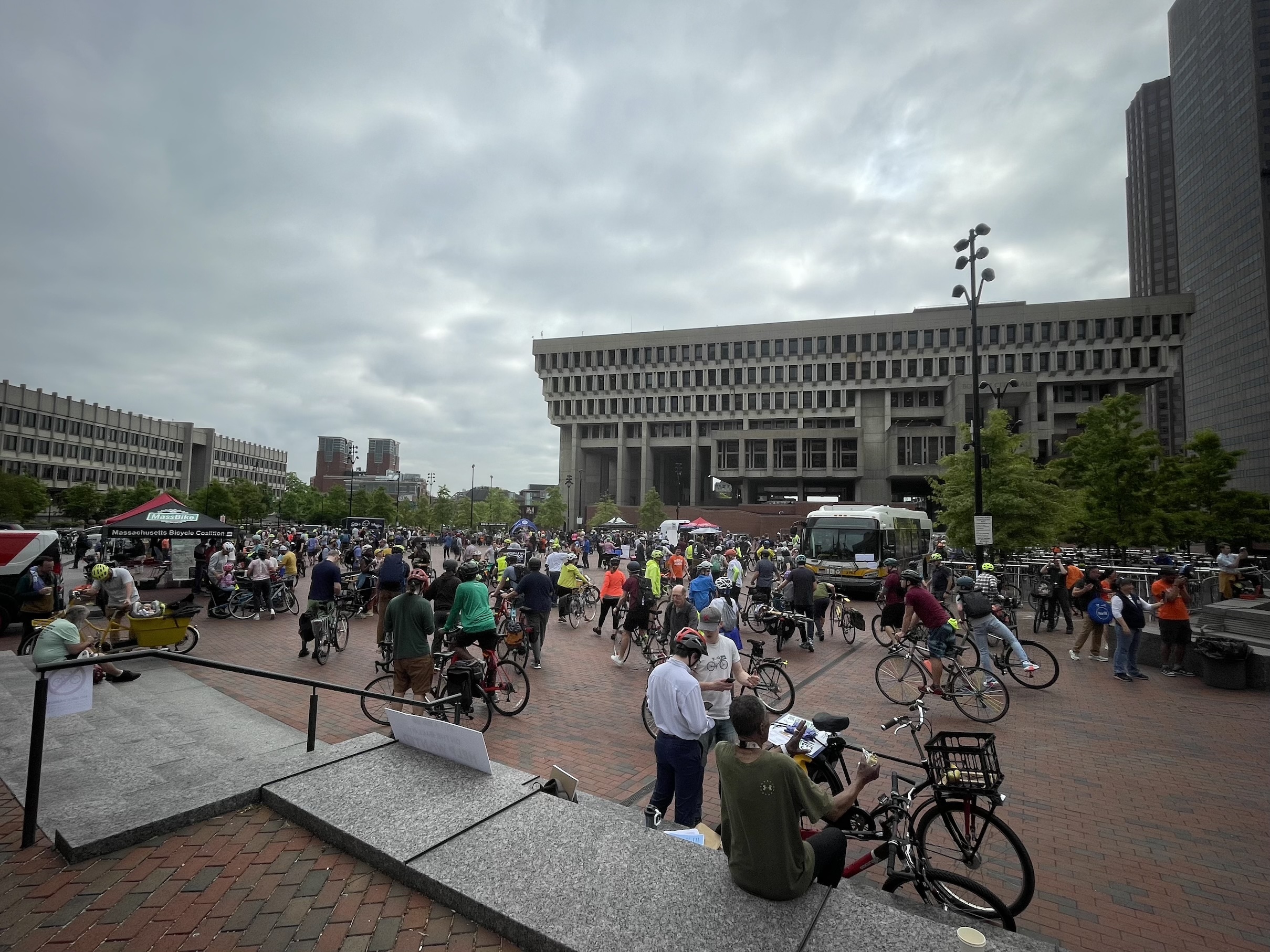 A crowd of people with bikes mingles in a large brick plaza with Boston's City Hall in the background at right.