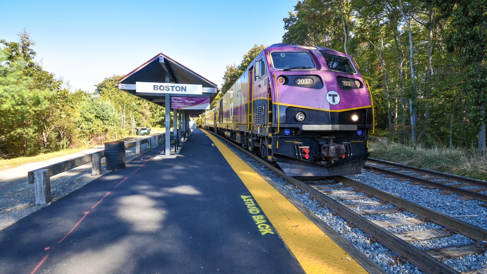 An MBTA commuter rail train alongside a asphalt-paved, low-level train station platform. In the middle distance is a small shelter over some benches on the platform with a sign reading "BOSTON".