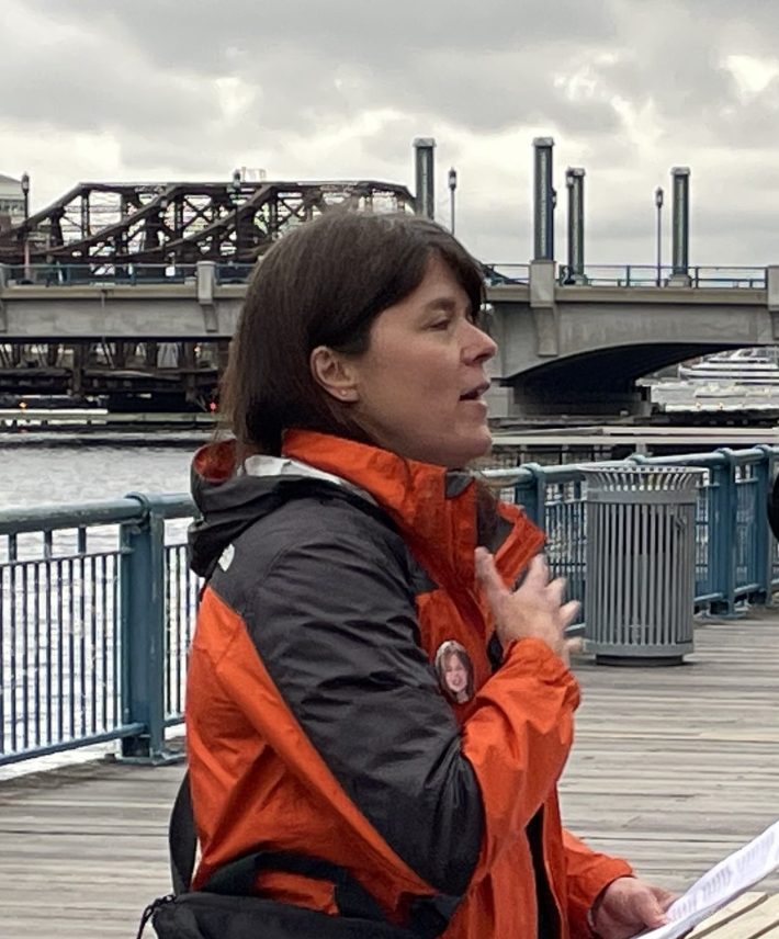 A woman in a red and black raincoat touches her hand to her chest while speaking next to the water on the Boston Harborwalk with the Moakley Bridge in the background.