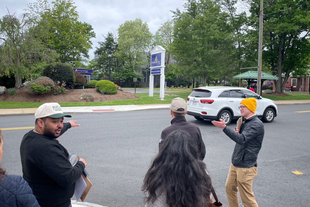 A man in a black jacket and yellow hat gestures at a city street next to three other people standing on a sidewalk.