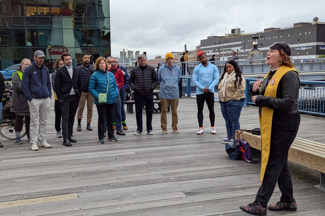 A woman wearing a yellow clerical stole over a leather jacket speaks in front of a small crowd of people on a city plaza.