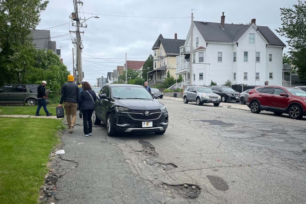 A city street with lots of potholes, lined with parked cars. Two pedestrians squeeze between a parked car and the edge of the asphalt at left.