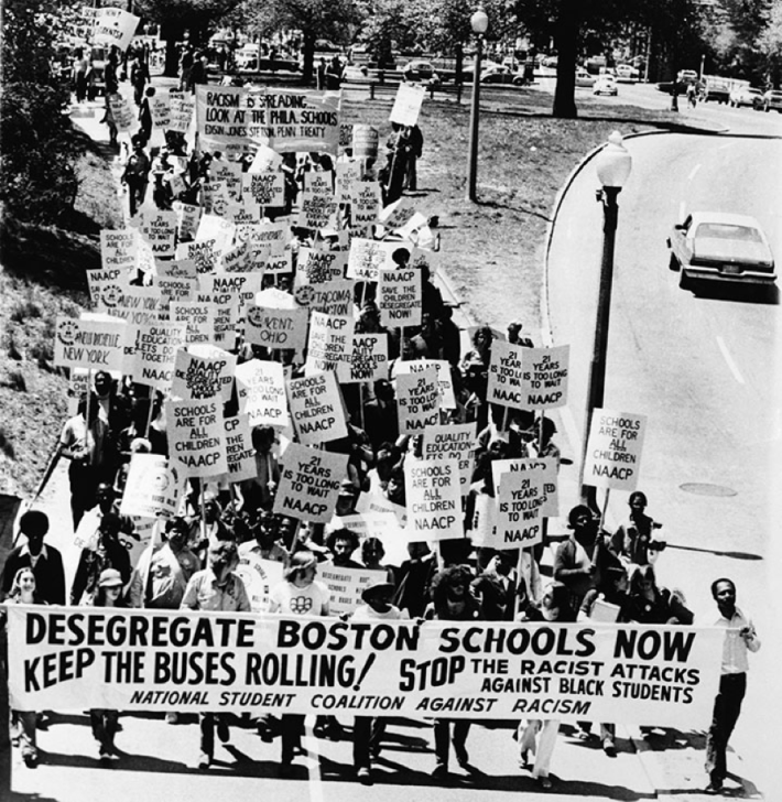 A large group of Black protesters march down a curved street holding signs calling for school desegregation and protection for Black students. A banner at the front reads “Desegregate Boston Schools Now – Keep the Buses Rolling! Stop the Racist Attacks Against Black Students.”