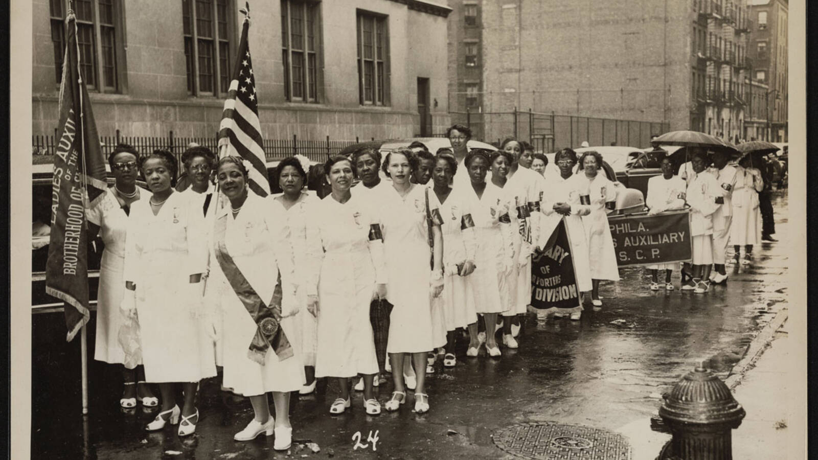 A group of Black women in white dresses standing on the street, holding banners for the Ladies Auxiliary of the Brotherhood of Sleeping Car Porters.