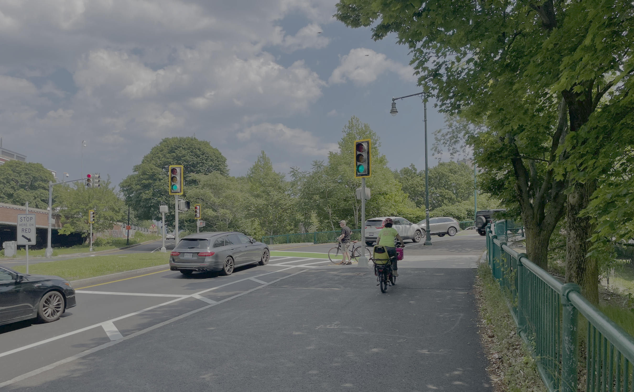 A person riding a bike along a ~14-foot wide bike path between some trees (at right) and a green railing and (at left) several cars passing through a traffic light at a small intersection. A second person is waiting to cross the crosswalk under the green traffic signal.