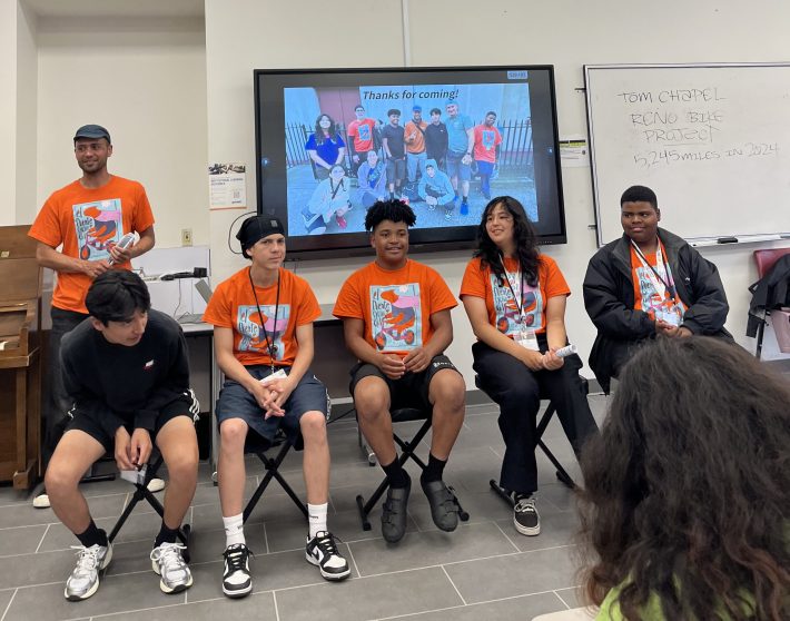 A panel of five youth wearing bright orange El Puente shirts sit in front of a classroom. A slide behind them says “Thanks for coming!” with a group photo.