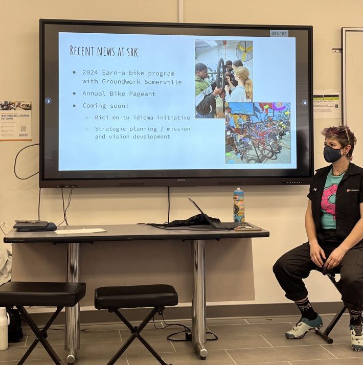 A short-haired woman in a mask sits next to a projector screen with the words "recent news at SBK: 2024 earn-a-bike program with Groundwork Somerville; Annual Bike Pageant; Coming soon: Bici en tu idioma initiative and Strategic planning"