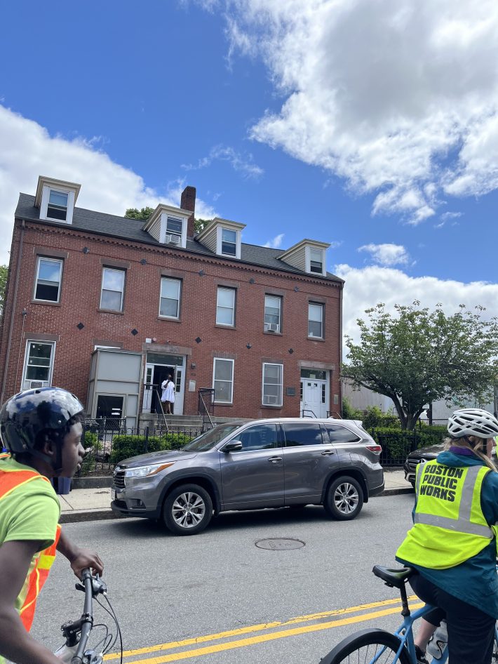 Riders pass a large red-brick residential building where a woman stands in the doorway looking on. Cyclists are wearing reflective vests and helmets.