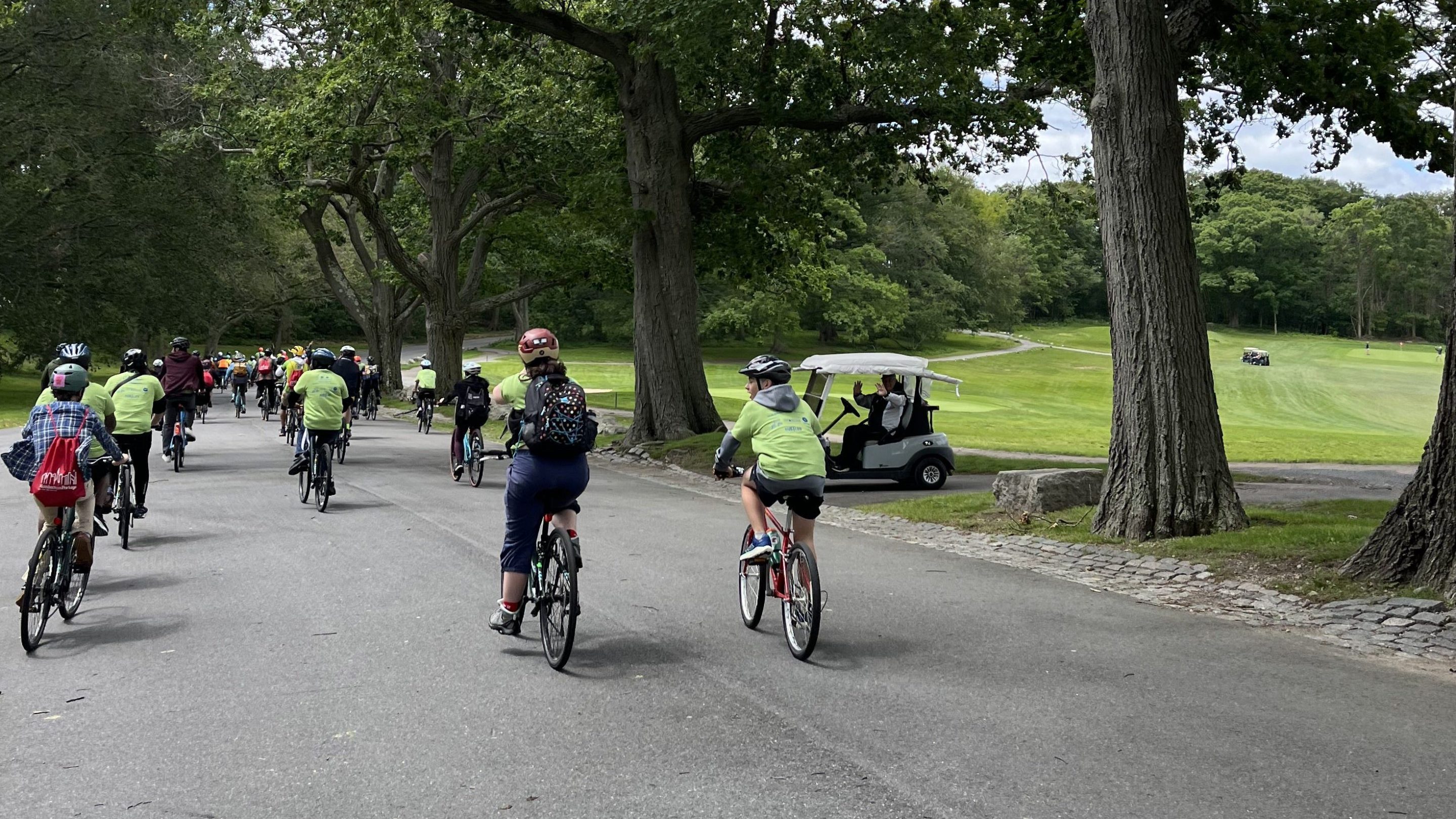 Dozens of cyclists in green Youth Bike Summit shirts ride past tall oak trees and manicured grass alongside a golf course, a man on a golf cart waves as the riders pass by.