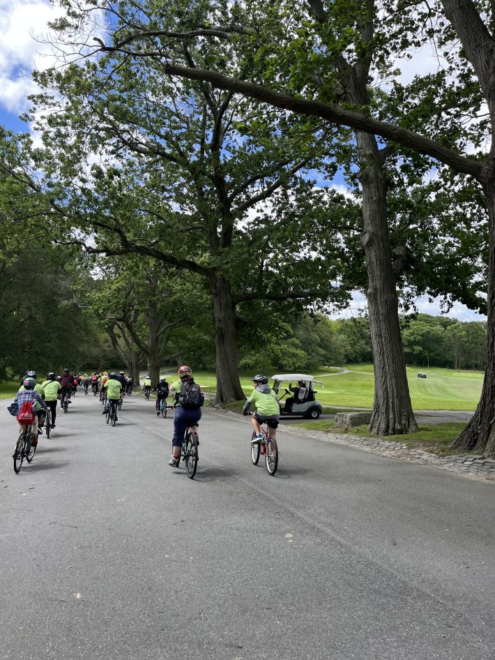 Dozens of cyclists in green Youth Bike Summit shirts ride past tall oak trees and manicured grass alongside a golf course, a man on a golf cart waves as the riders pass by.