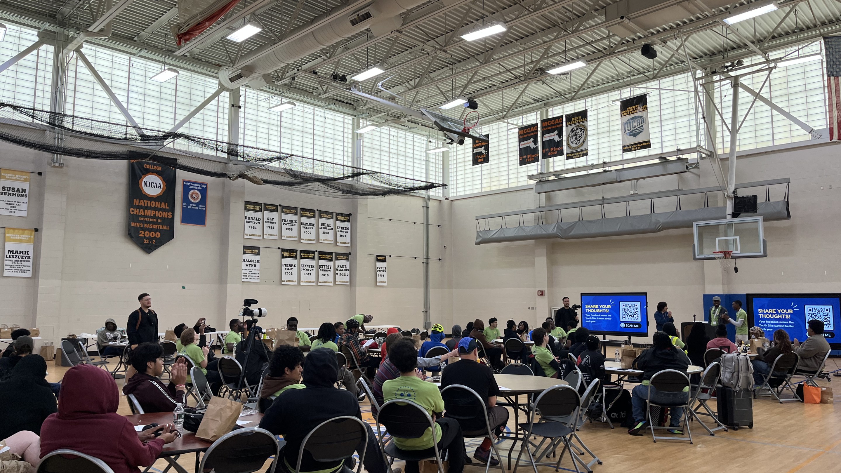 Participants wearing green Youth Bike Summit shirts gather in a gymnasium for a group session, sitting at round tables in front of presentation screens.