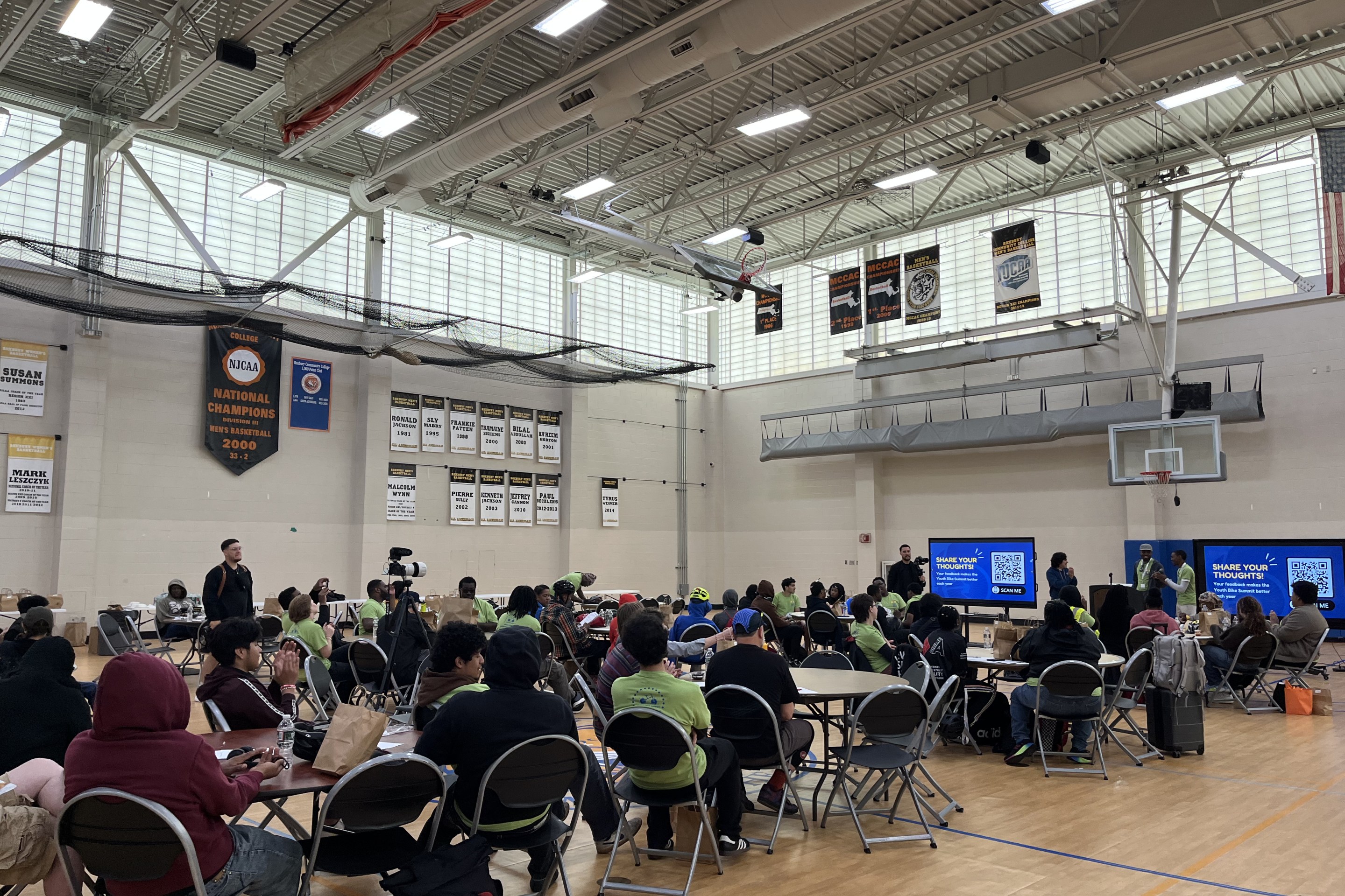 Participants wearing green Youth Bike Summit shirts gather in a gymnasium for a group session, sitting at round tables in front of presentation screens.