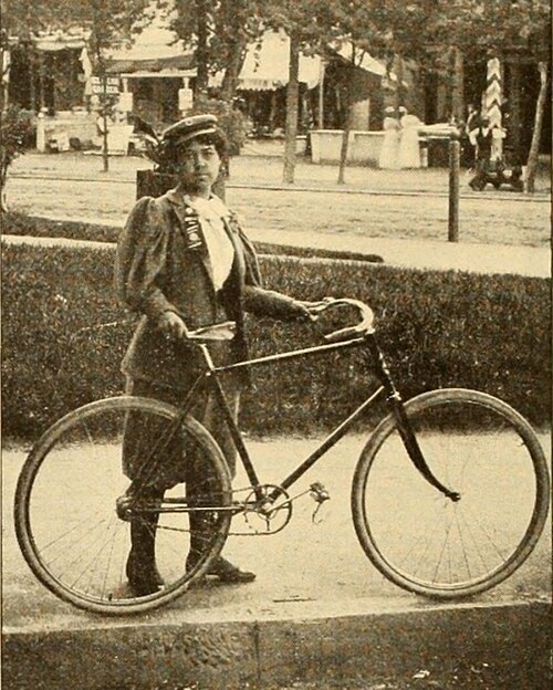 A young Black woman in a dress stands beside a bicycle on a city street, looking directly at the camera.