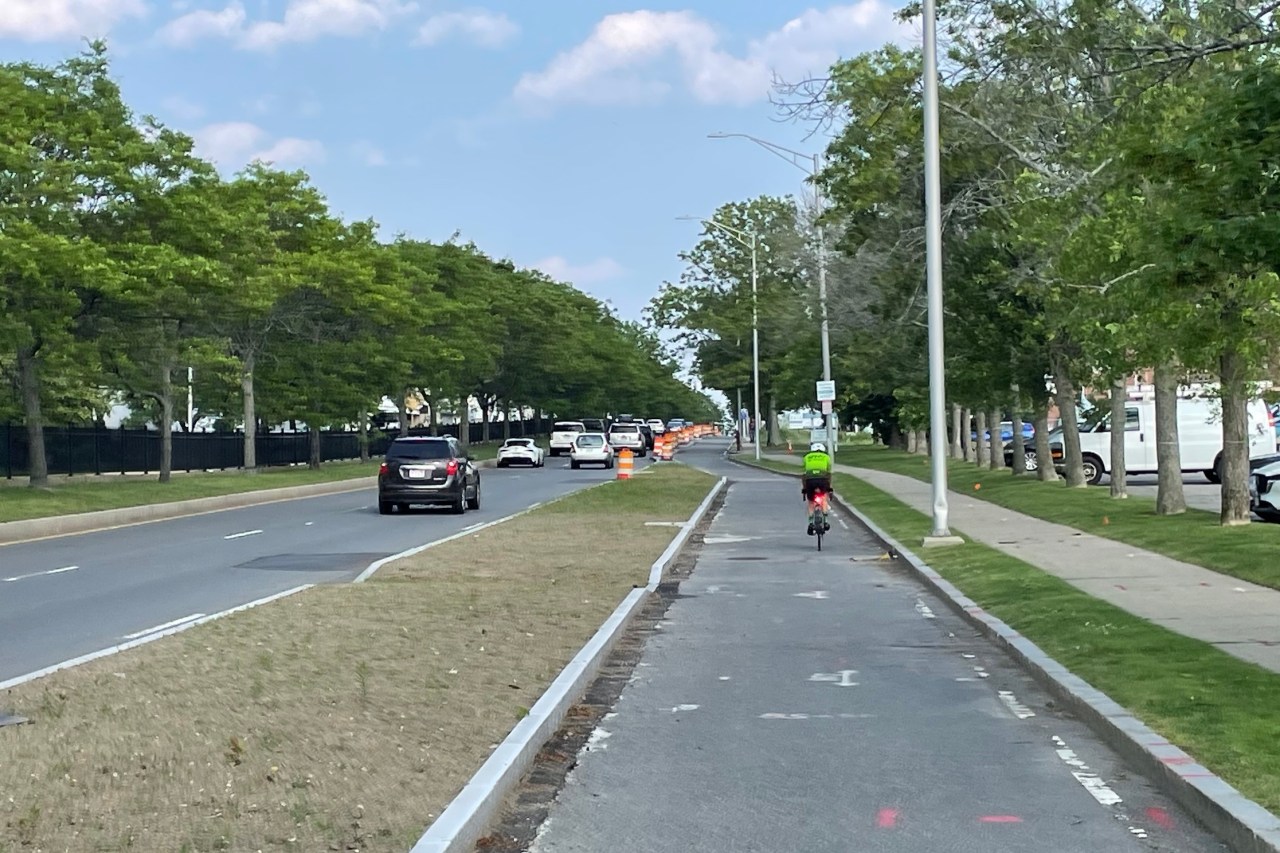 A man on a road bike rides along a paved path between a highway on the left and a sidewalk lined with trees on the right. Construction barrels line the path into the distance.