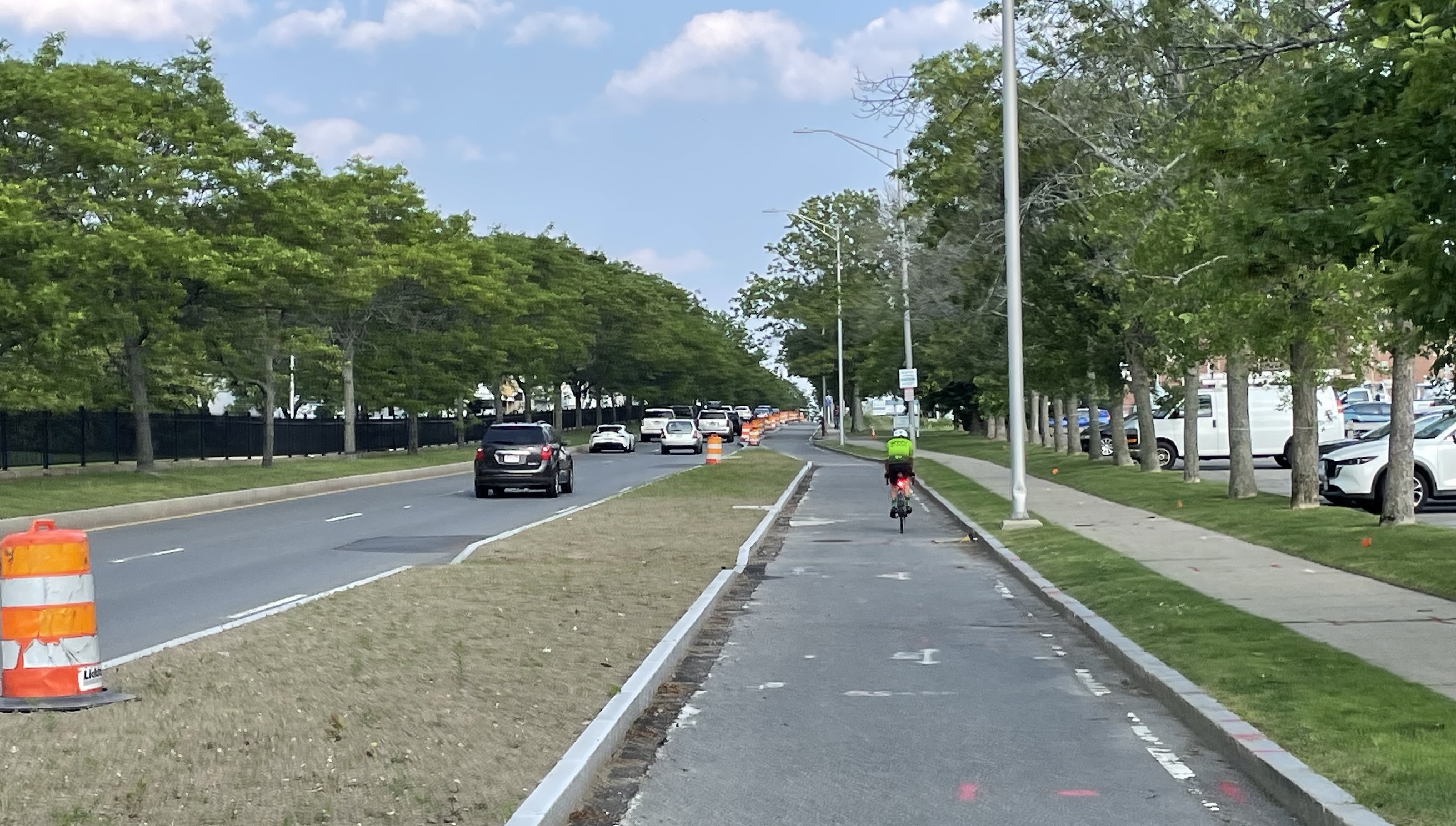 A man on a road bike rides along a paved path between a highway on the left and a sidewalk lined with trees on the right. Construction barrels line the path into the distance.