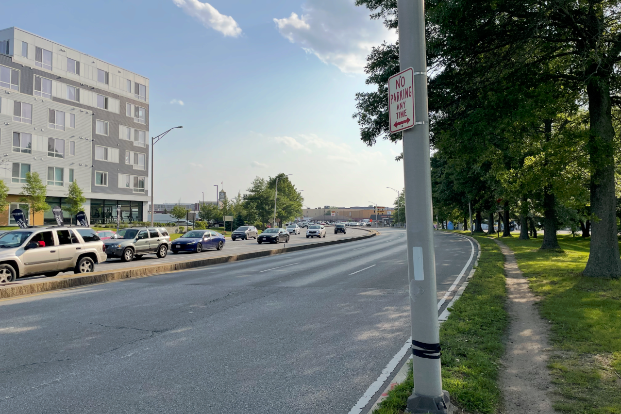 A well-worn dirt path runs alongside an allee of trees next to a large 6-lane-wide divided highway. On the left edge of the photo, on the opposite side of the roadway, is a 6-story apartment building. A metal street lamp pole in the foreground bears a "no parking any time" sign.