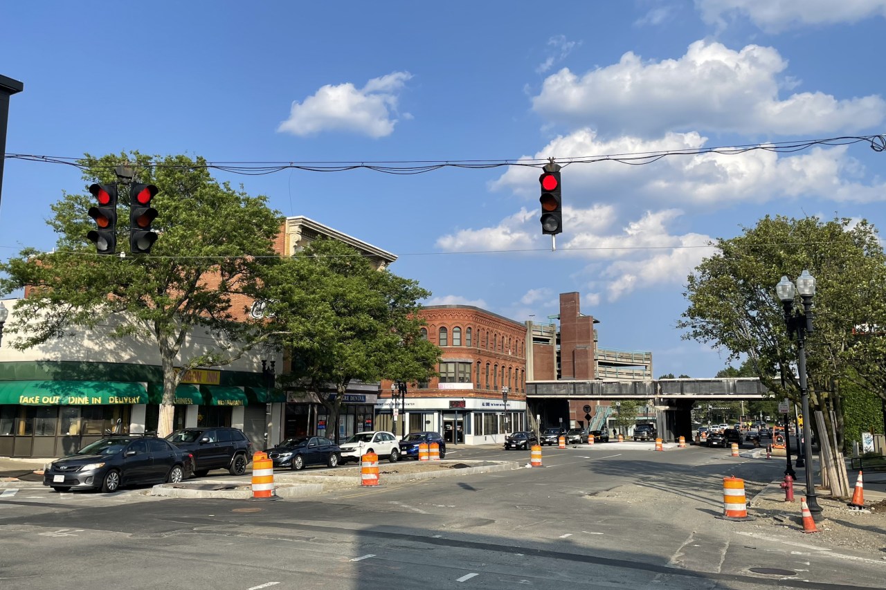 A city intersection under construction with several orange cones and construction barrels. On the opposite side of the intersection are several multi-story brick buildings and in the distance is a railroad bridge over the street.
