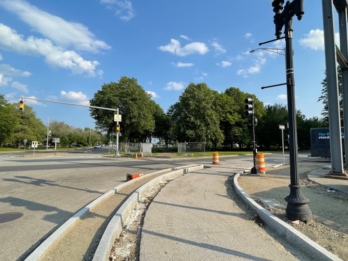 A bike path under construction alongside a wide multi-lane intersection. Several construction barrels and cones line along the edges of the road. On the other side of the intersection is a grassy parkland with mature deciduous trees, and a traffic light.