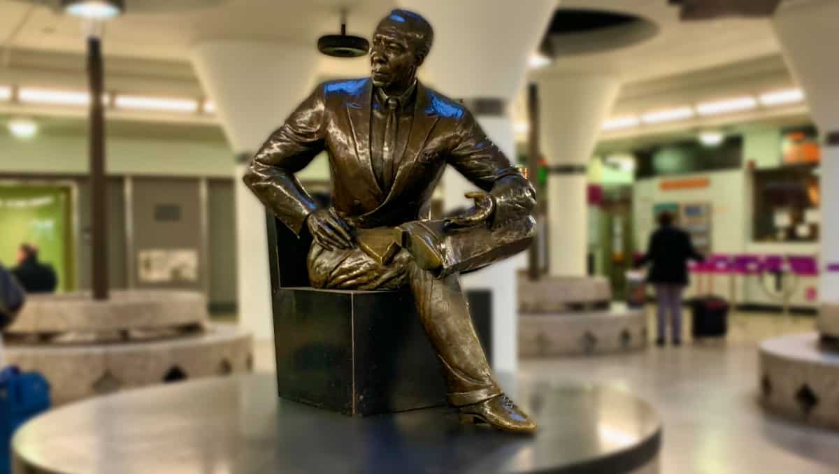 A bronze statue of A. Philip Randolph seated with one leg crossed over the other, holding a book. This statue is located inside Boston’s Back Bay Station.