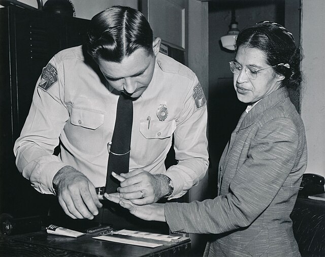 Rosa Parks, wearing a suit and glasses, looks down as a white deputy sheriff fingerprints her during booking in 1956.