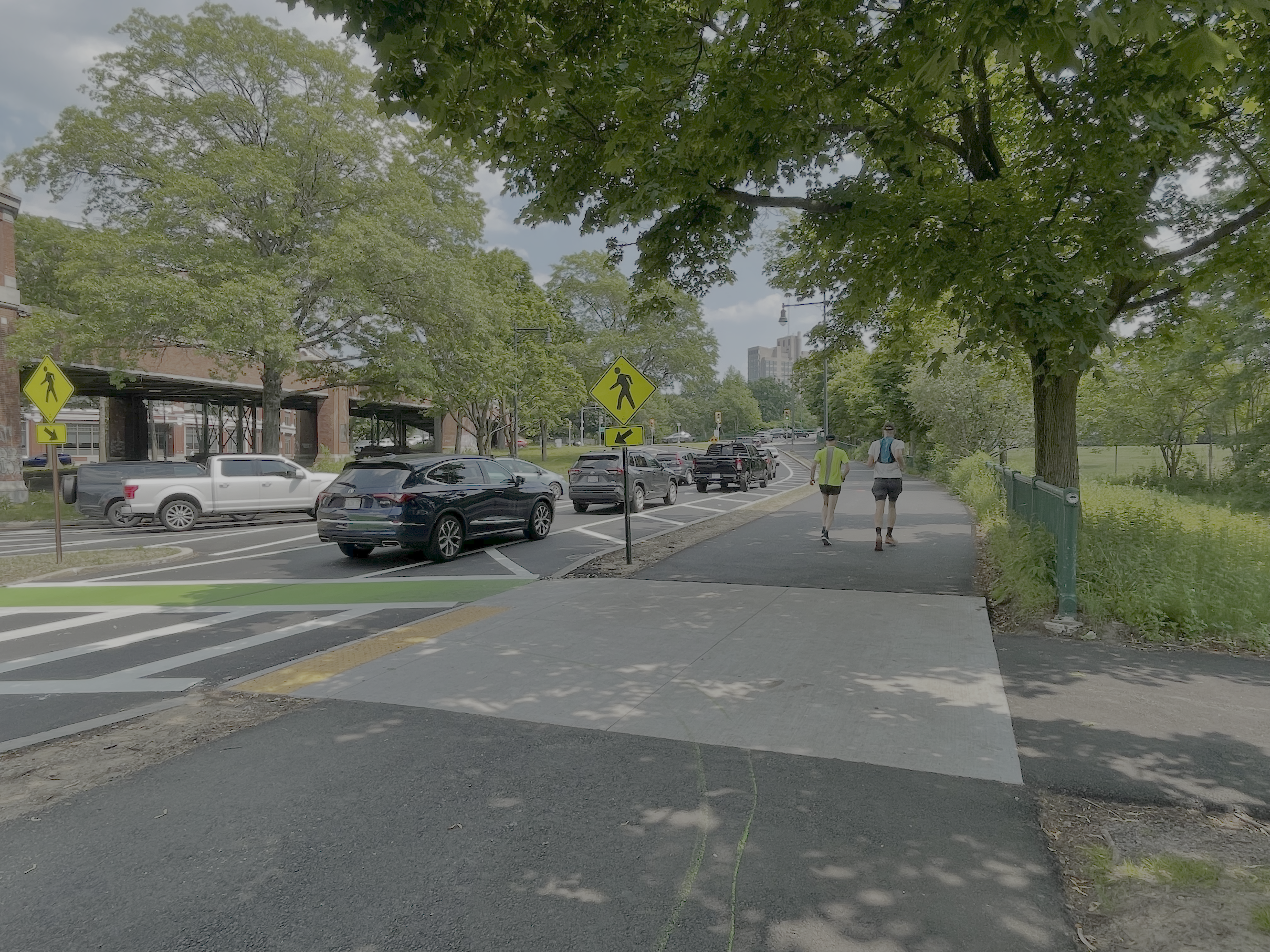 A wide sidewalk path runs alongside a line of cars waiting for a traffic signal in the distance. To the right is an overgrown meadow and a large leafy tree that shades the path. On the left edge a crosswalk leads across the roadway. In the middle distance to the left is a brick-clad highway viaduct.