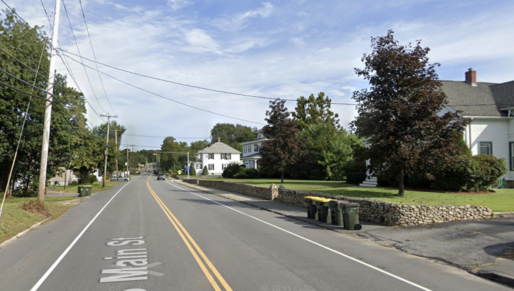 A two-lane roadway with a double-yellow stripe down the center runs through a suburban neighborhood