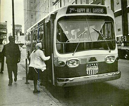 A woman boards a city bus in Winston-Salem, North Carolina. The bus sign reads "3rd - Happy Hill Gardens".