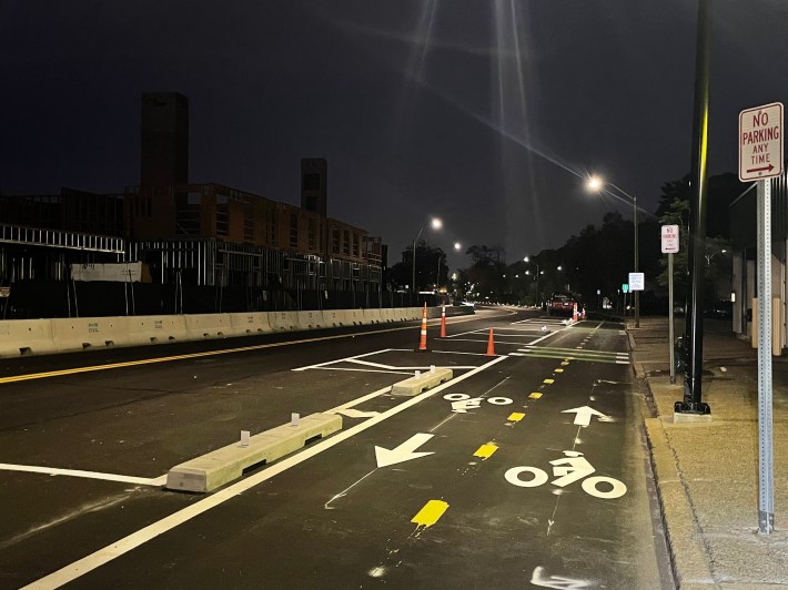A city street pictured in the evening after sundown. In the foreground is a two-way bike path, separated from the rest of the street by concrete curb barriers. On the opposite side of the street is a construction site.