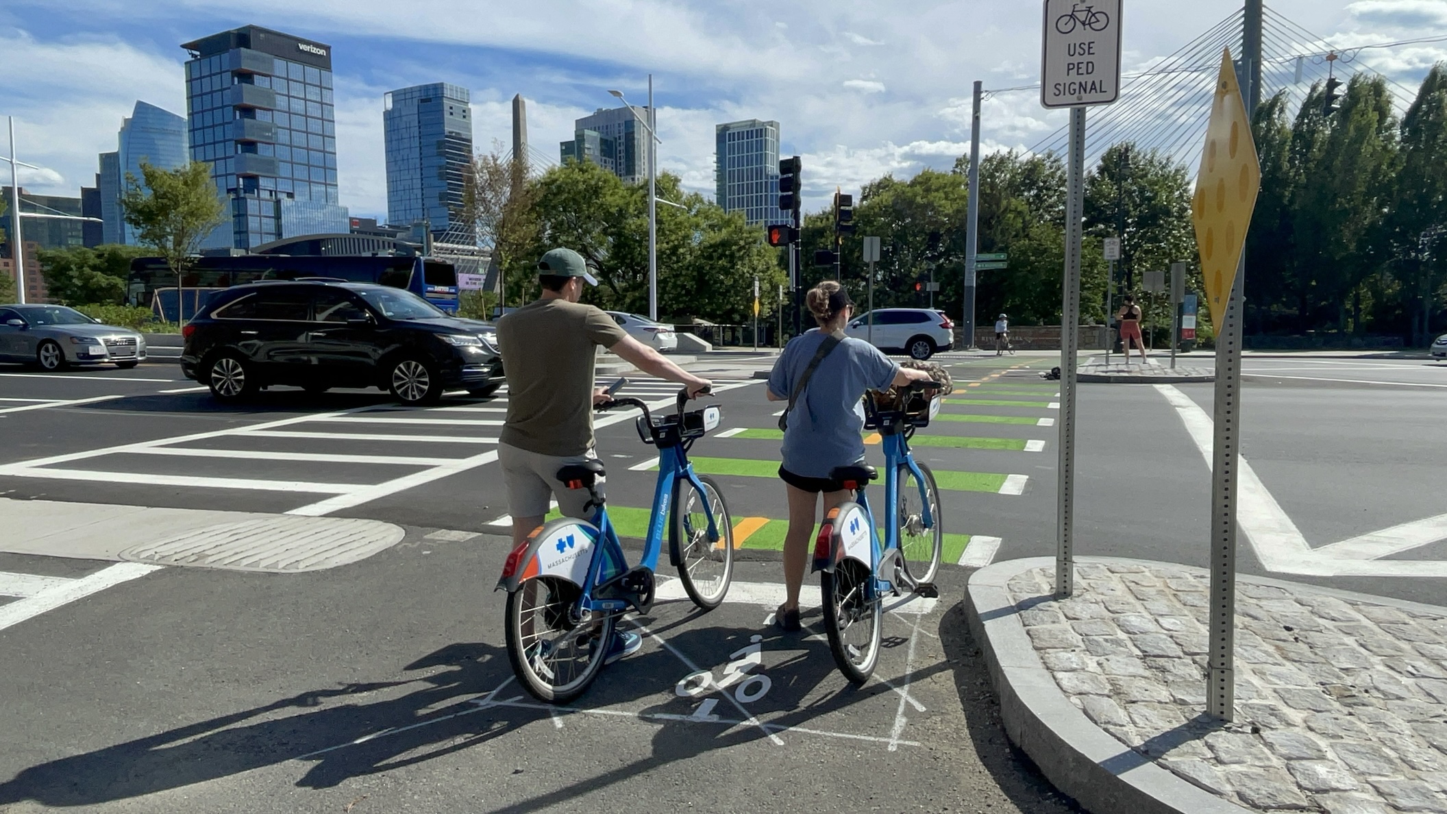 Two people with Bluebikes wait to cross a wide street towards a park on the other side. On the horizon to the left are four glass towers in downtown Boston.