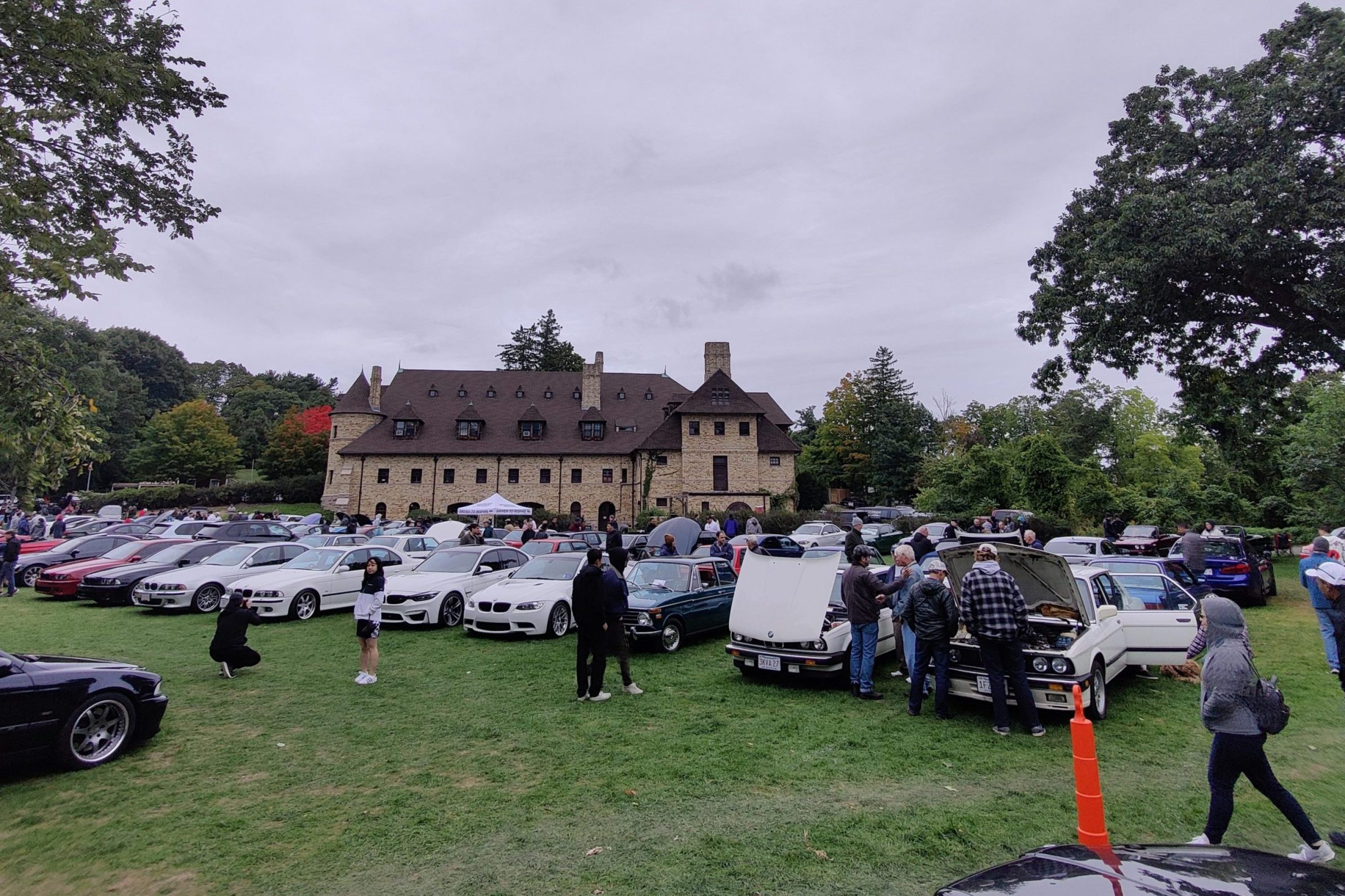 People mingle around rows of parked cars on a broad lawn in front of a Romanesque Revial-style stone building with many dormers on its roof.