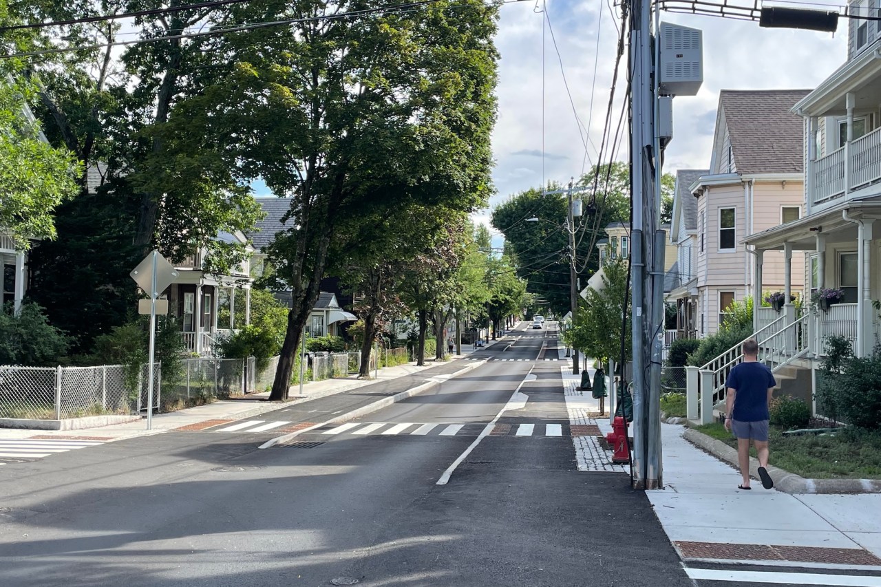 A city street lined with small 3-story multi-family residential buildings. Two concrete curbs in the middle of the street divide a single lane of motor vehicle traffic from two sidewalk-level bike lanes on either side. White zebra-striped crosswalks cross the street every block into the distance. The street is also lined with leafy trees of varying sizes and ages, some of which are very large and mature, others of which have just been planted.