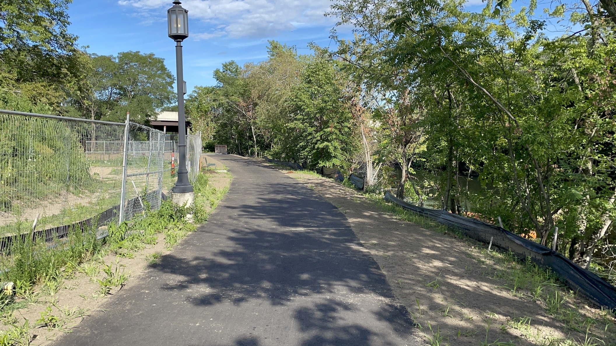 A recently-paved bike trail runs along a riverbank (right) next to a chain link construction fence and a street lamp (at left). In the distance a highway overpass is visible through the trees.