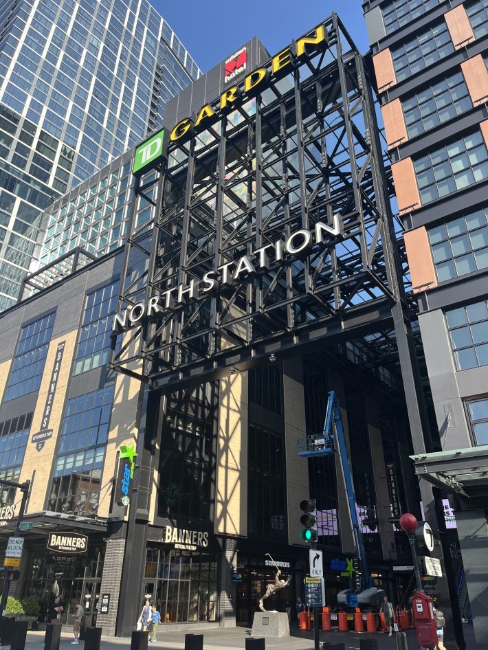 The North Station entrance on Causeway Street underneath a sign for TD Garden, bookended by tall buildings and  busy storefronts.