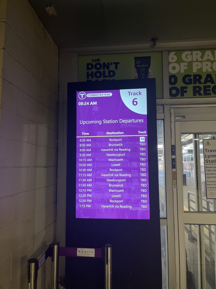 A digital screen showing MBTA commuter rail departures including times for Rockport, Newburyport, and Wachusett, next to a sign that says "Track 6."
