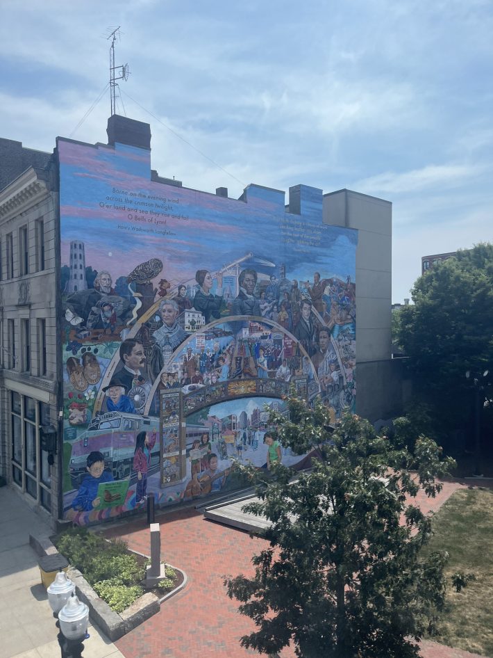A large mural on a building in Lynn, MA showing historical and cultural figures and community scenes. The top of the mural features a quote by Henry Wadsworth Longfellow.