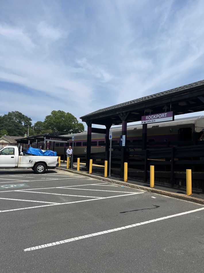 A view of the Rockport MBTA Commuter Rail platform with a train at the station and a few people waiting nearby.