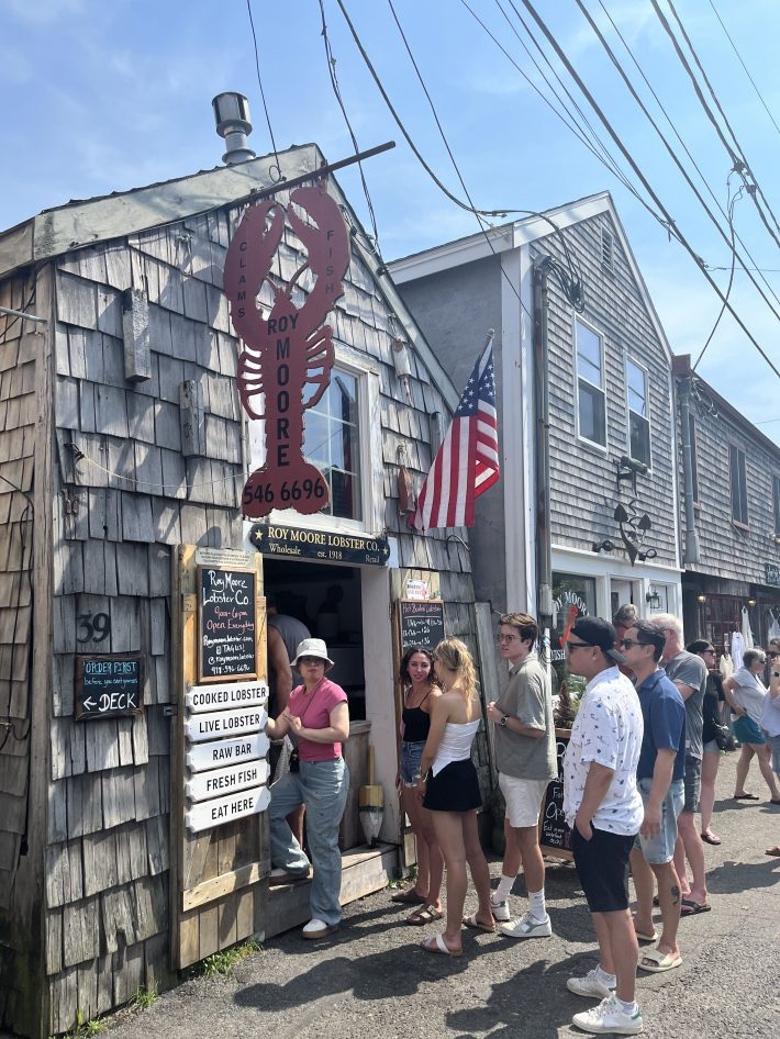 A line of people wait to order at Roy Moore Lobster Co., a small shack with signs for lobster, a raw bar, and fresh fish.