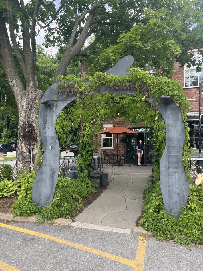 A metal archway covered in greenery reading "Foam Brewers" leads to a patio with metal chairs and tables outside a brick building.