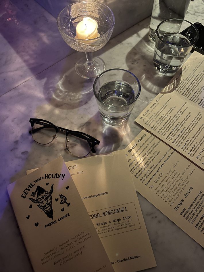 A dimly lit cocktail bar with a marble surface, a glass holding a lit candle, two glasses of water, black-rimmed glasses, and a drink menu.