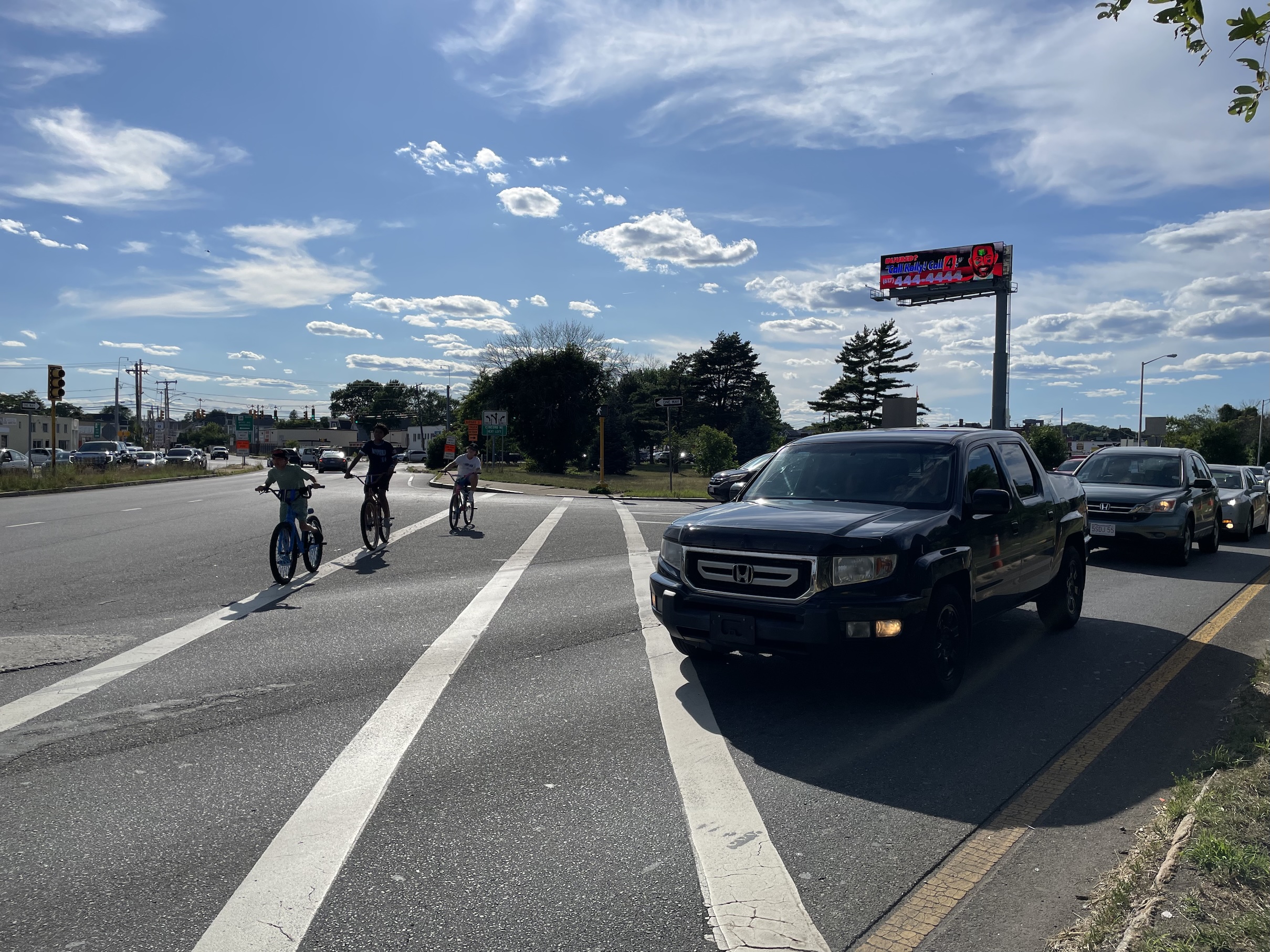 A line of large cars waits at a highway off-ramp at right as three kids on bikes approach the camera on a long crosswalk next to a multi-lane highway. Above the offramp at right is a large electronic billboard rising up into the blue sky.