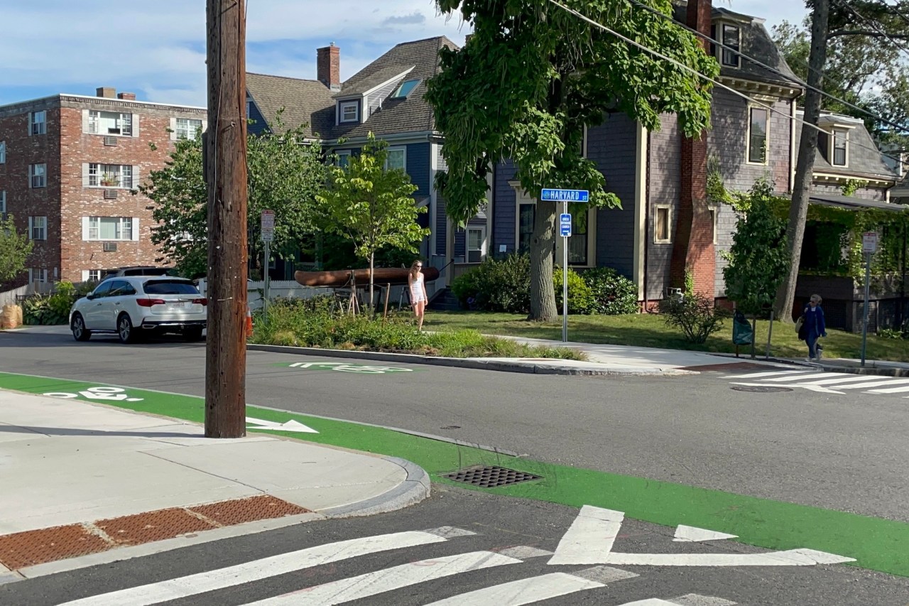 A residential city street lined with multi-story apartment buildings. A blue street sign in the center of the photo at an intersection on the opposite side of the street says "HARVARD ST." In the foreground a green-painted bike lane runs along the near side of the street from the left edge of the photo to the lower right corner. A garden with trees and shrubs lies between the street and the sidewalk on the opposite side of the street.