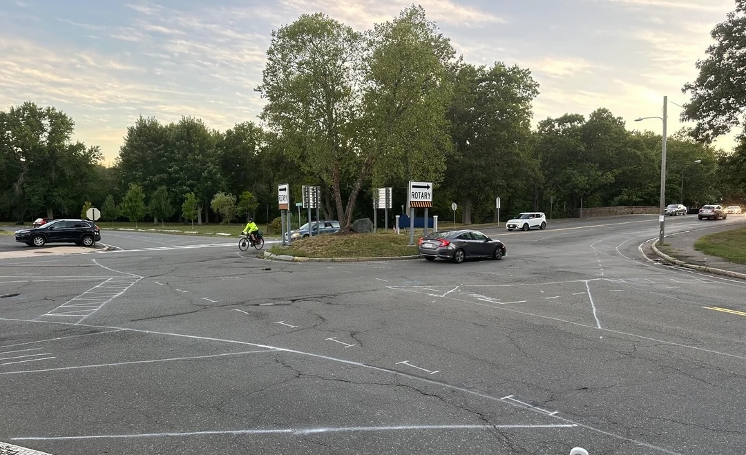 A street-level photo of a wide traffic circle with a small island in the middle with a tree and signs that read "ROTARY" above black-and-yellow stripes. Spray-painted markings in the roadway indicate the future locations of new street lane markings.