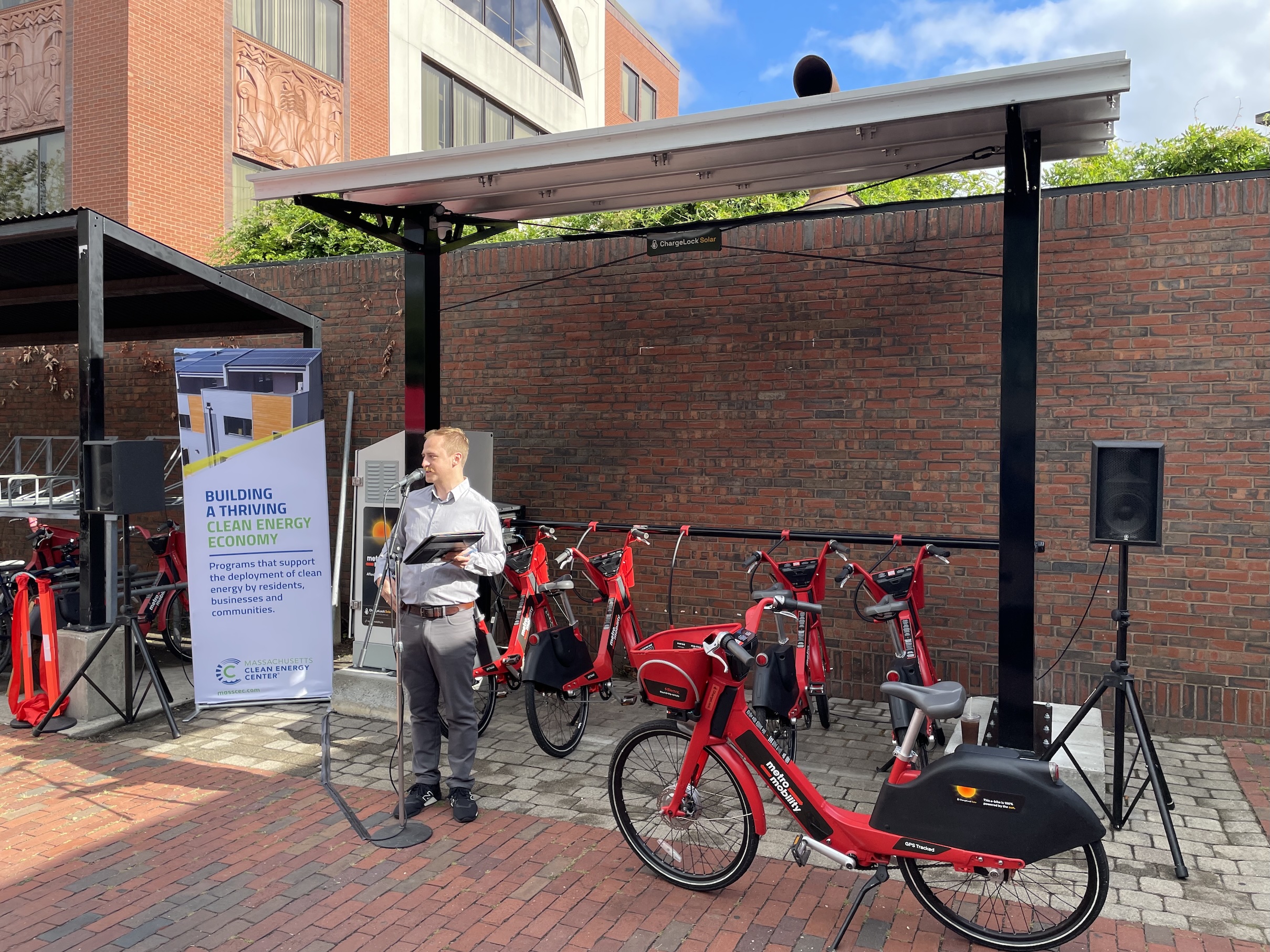 A man wearing a collared shirt speaks at a microphone in front of a bike rack with four red e-bikes. An additional red bike with the words "metro mobility" on its frame rests on its kickstand in front of the bike rack. Above the bike rack is a rain shelter mounted with solar panels.