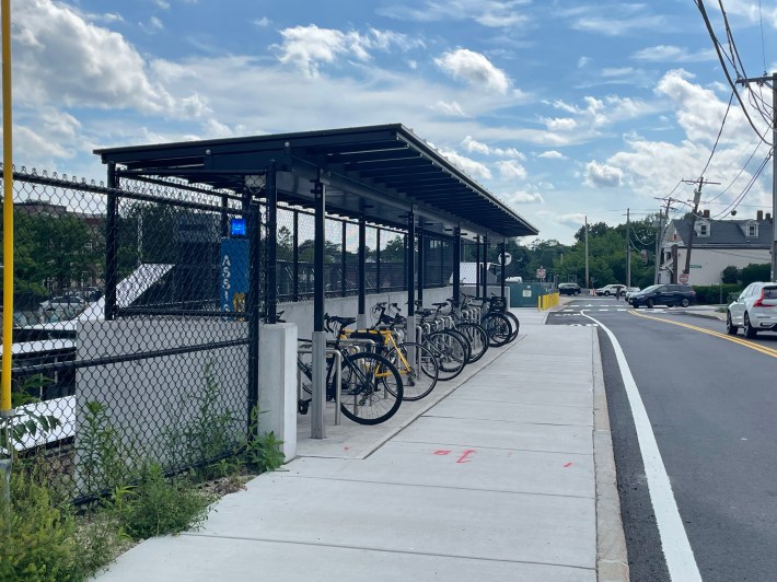 A bike rack sheltered under a metal roof, next to a sidewalk.