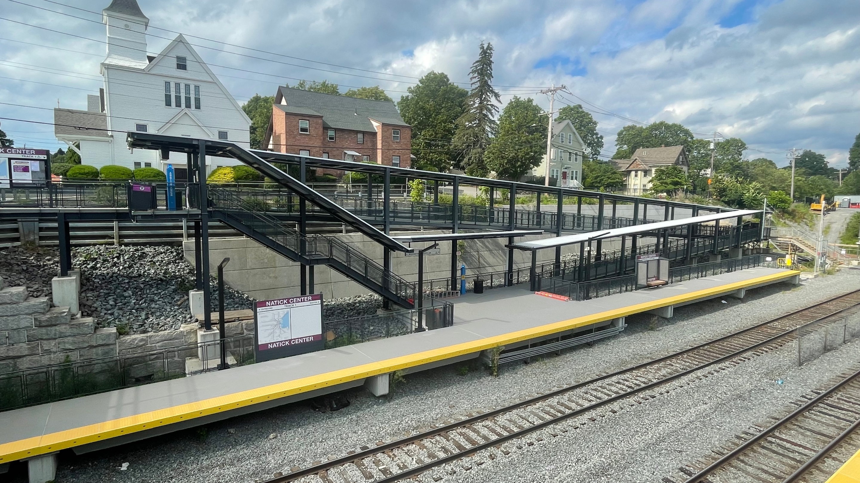 An elevated view of a commuter rail station across two railroad tracks. A sign on the platform reads "NATICK CENTER"