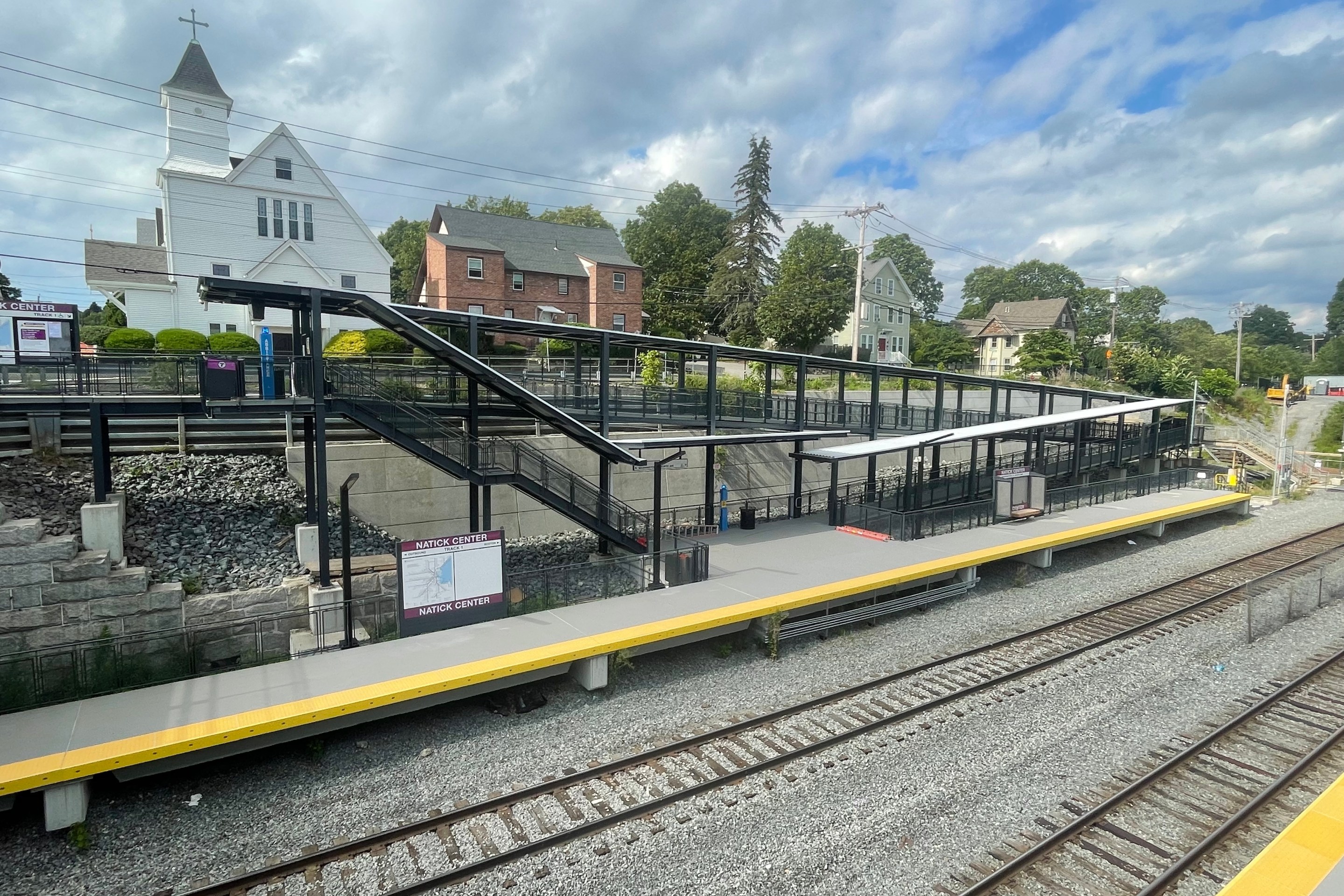 An elevated view of a commuter rail station across two railroad tracks. A sign on the platform reads "NATICK CENTER"