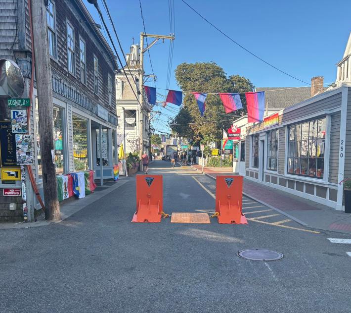 Two bright-orange metal barriers chained to each other block access to a narrow street lined with shops.
