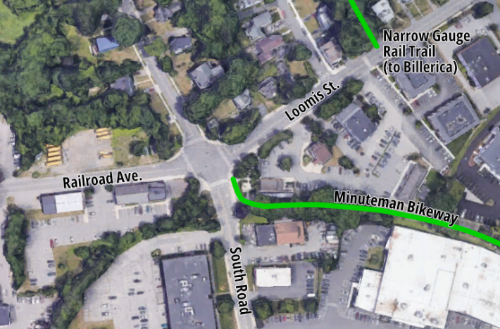Satellite map of an intersection surrounded by homes to the north and warehouses and parking lots to the south. A green line from the center to the right edge is labelled "Minuteman Bikeway". The north-south street through the intersection is labelled South Road. The street to the left of the intersection is labelled Railroad Ave., and the street to the right is labelled Loomis St.