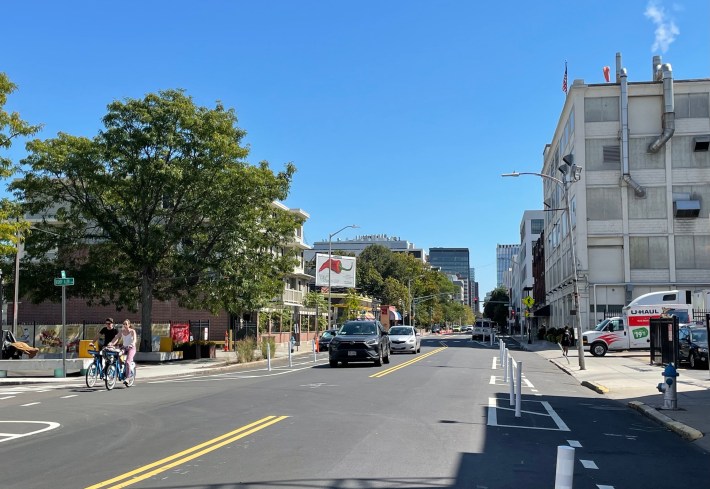 Two people on bikes ride down a protected bike lane on a city street lined with 4-5 story older industrial buildings and trees.